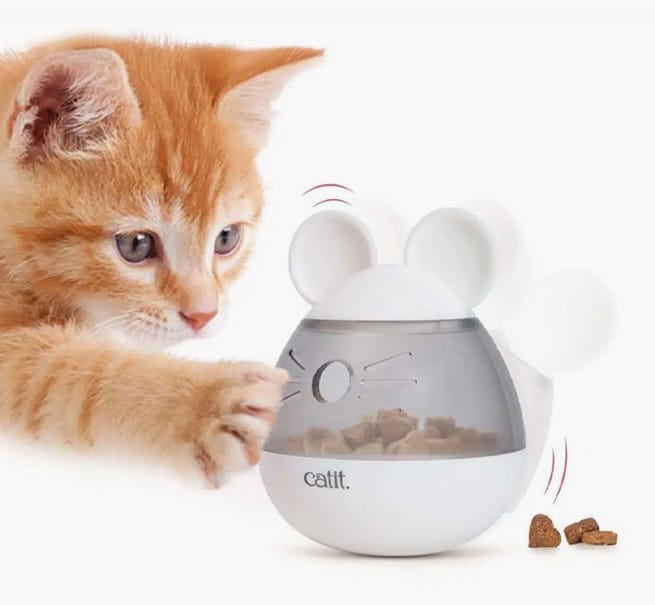 An orange tabby kitten batting at a white, mouse-shaped Catit PIXI treat dispenser that is releasing dry food kibble