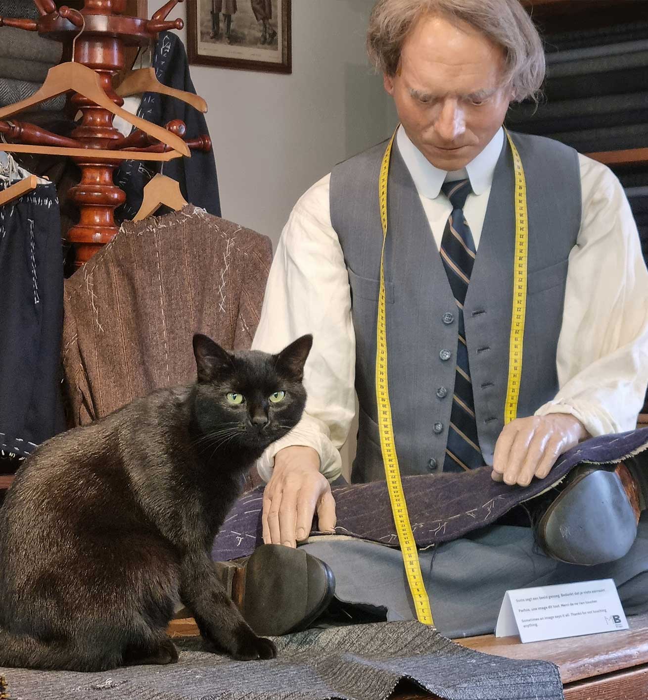 A black cat sits next to a mannequin of a tailor working on a piece of fabric in a workshop