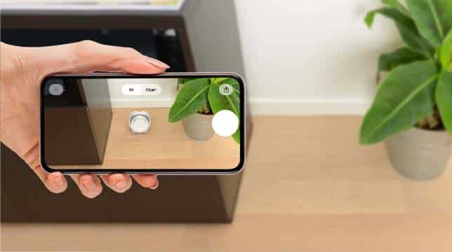 A hand holds a smartphone displaying an augmented reality view of a Catit PIXI litter box in a room next to a plant
