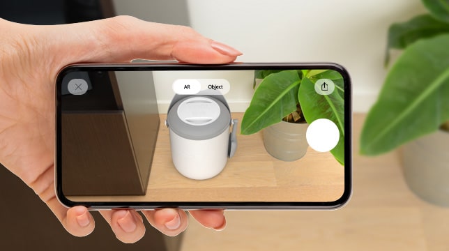 A hand holds a smartphone displaying a PIXI vacuum in an augmented reality view on a floor next to a potted plant