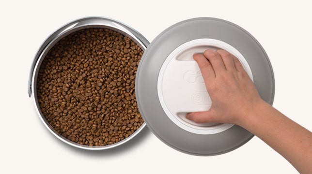 A hand opening the lid of a Catit PIXI vacuum food container filled with dry cat food