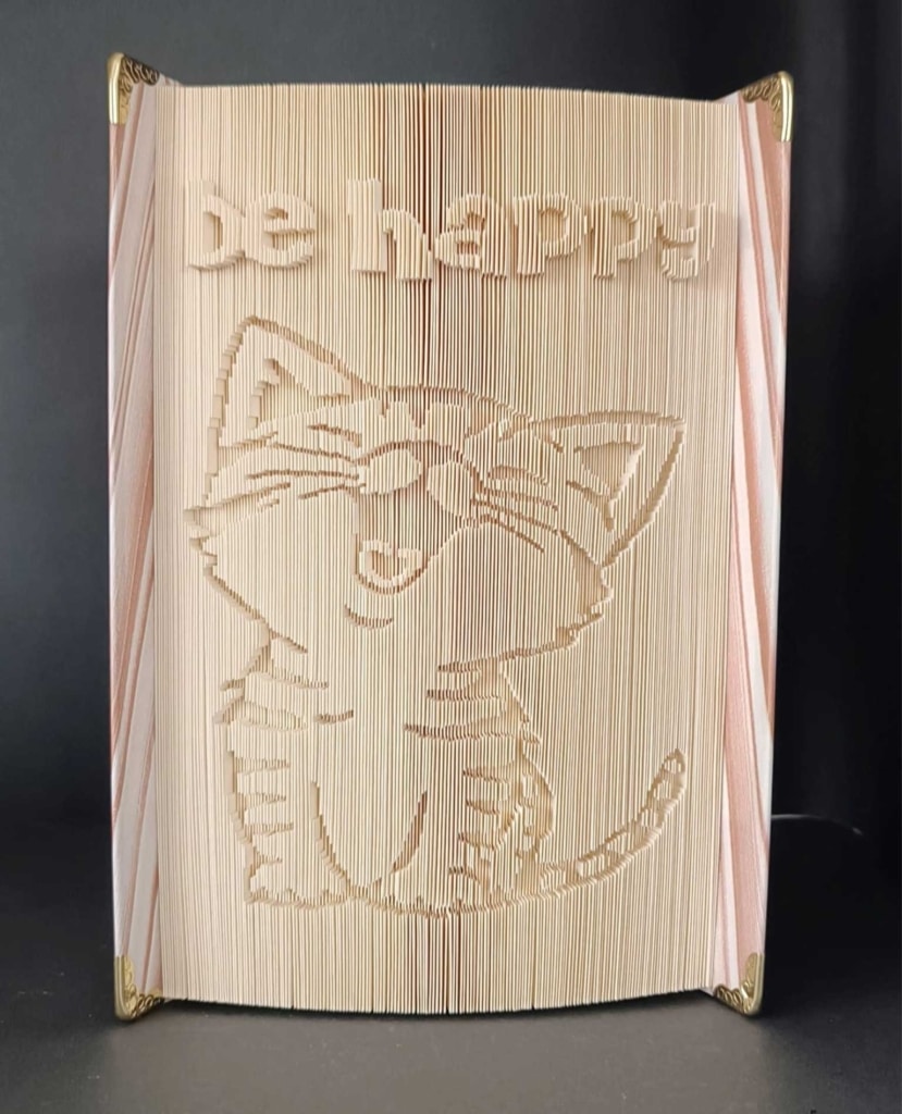 Book art featuring a folded paper sculpture of a cat with the words be happy
