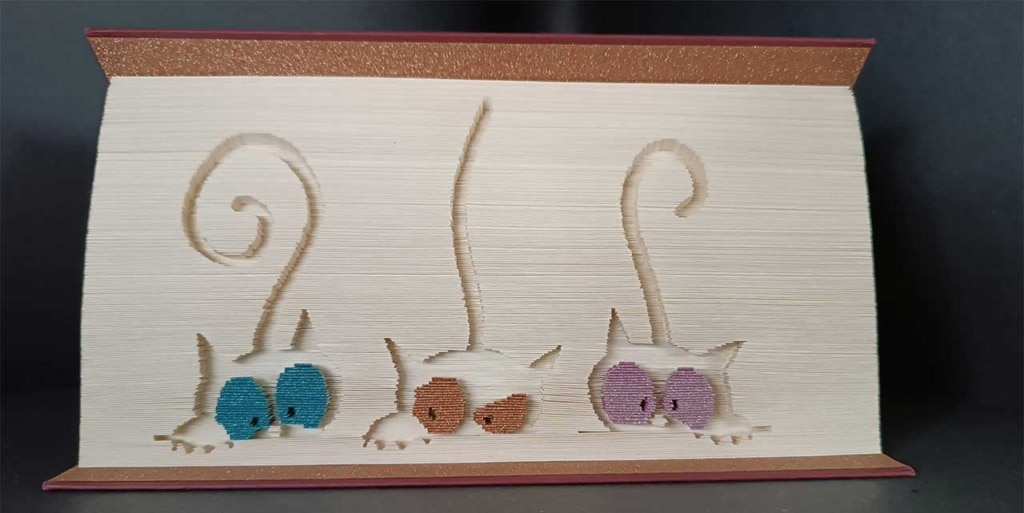 Book art sculpture featuring three cats with colorful eyes peeking over the pages