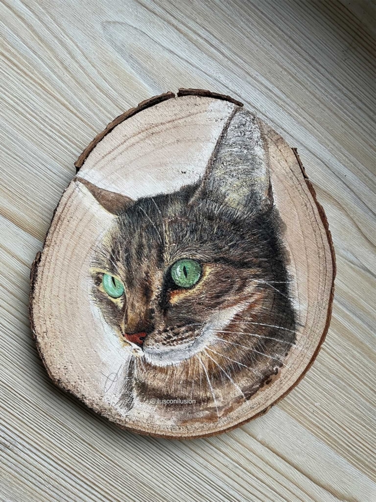 A detailed portrait of a tabby cat with bright green eyes painted onto a circular slice of natural wood
