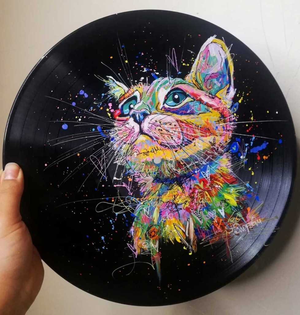 A hand holds a vinyl record painted with a colorful, abstract portrait of a cat looking upward