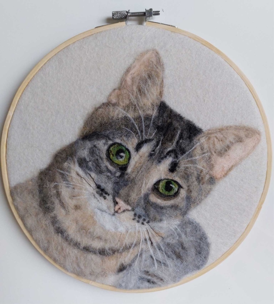Needle-felted portrait of a tabby cat with green eyes, displayed in a circular wooden embroidery hoop