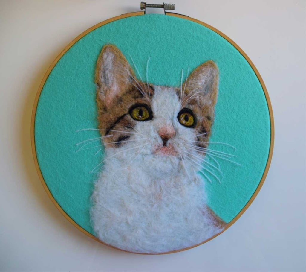 Needle-felted portrait of a brown and white cat in a wooden embroidery hoop against a teal background