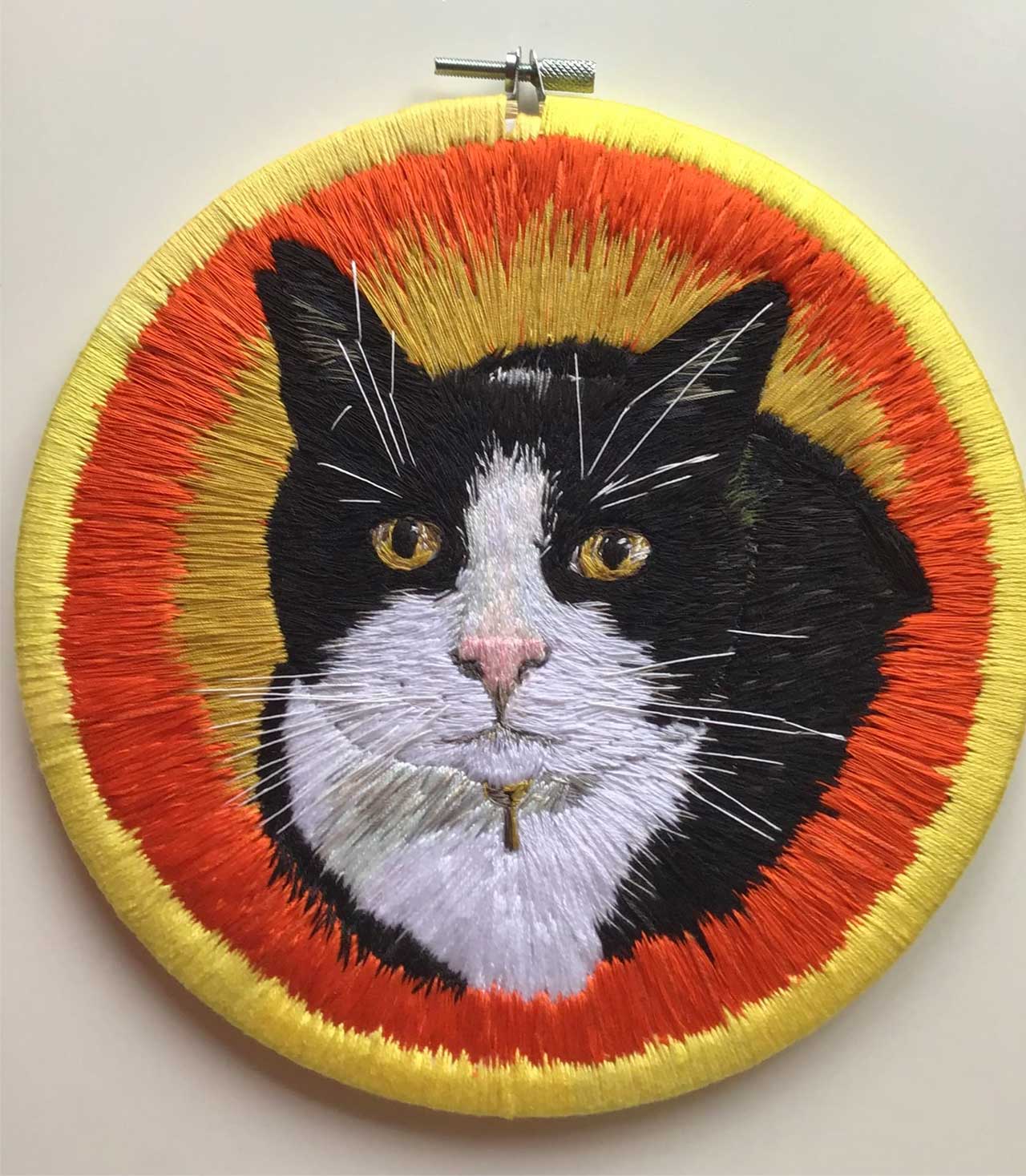 An embroidery hoop featuring a portrait of a black and white tuxedo cat against a vibrant yellow and orange sunburst background