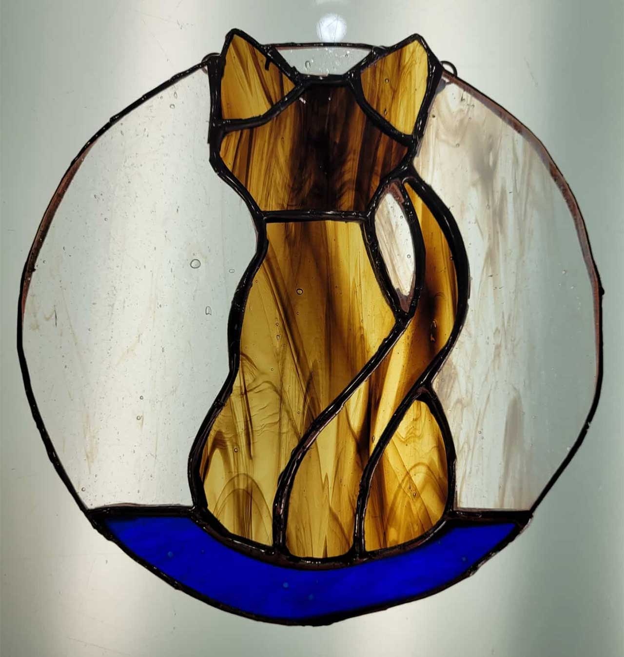 Stained glass art of a brown cat sitting against a circular white background with a blue base