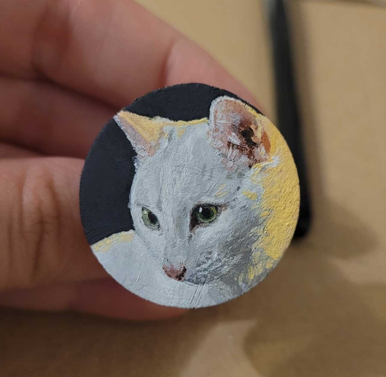 A small, circular hand-painted portrait of a white cat with green eyes held between fingers