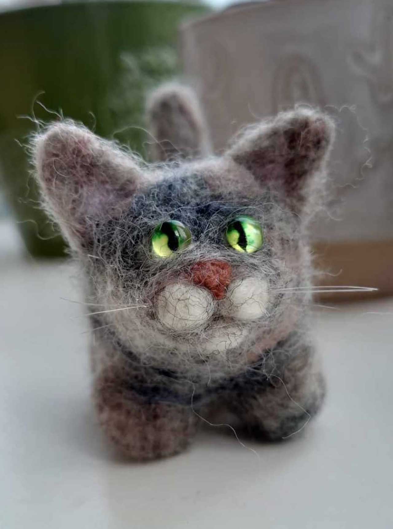 A small, handmade needle-felted cat figurine with grey fur and bright green eyes