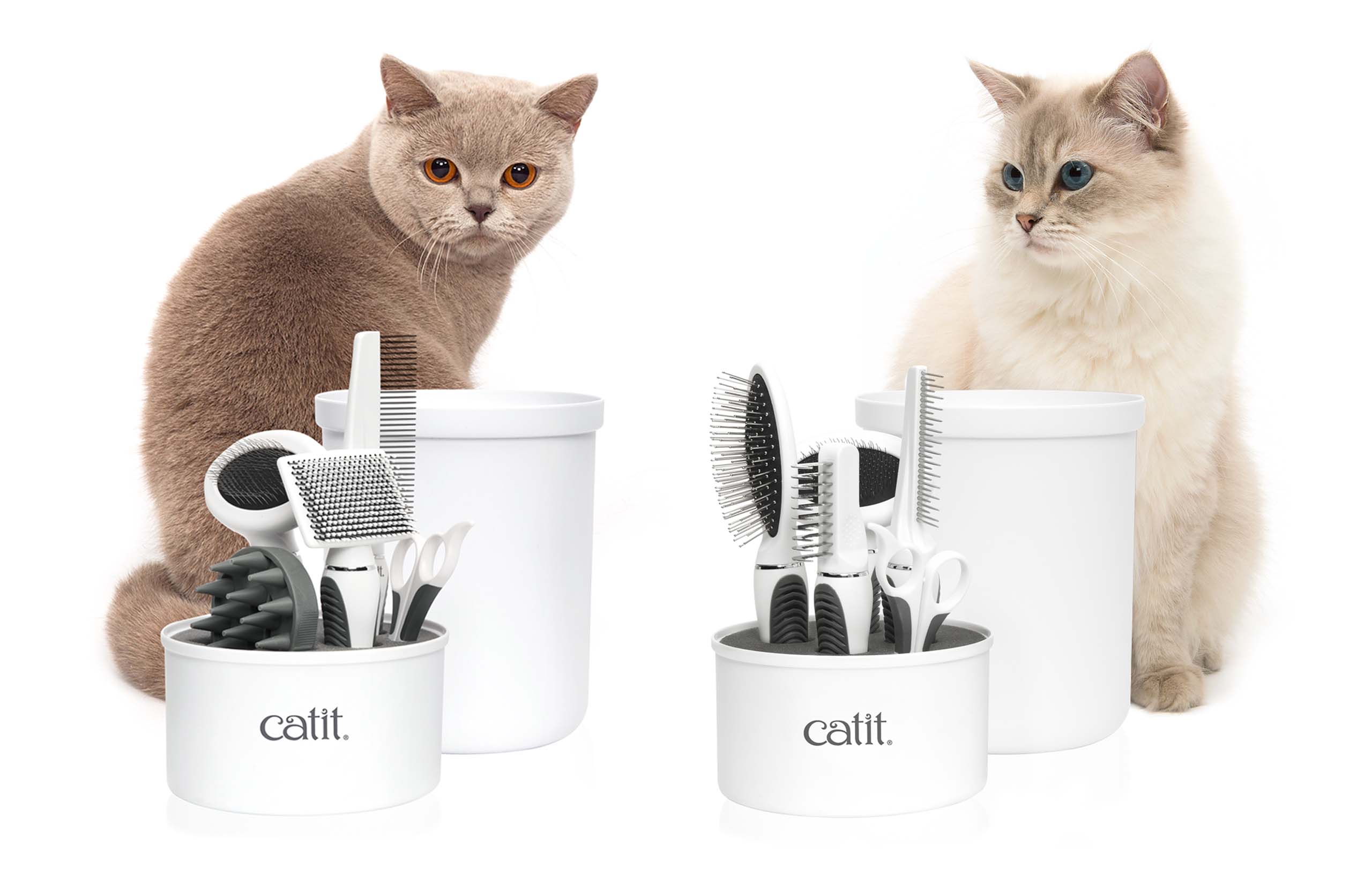 Two cats sitting behind Catit grooming kits containing various brushes, combs, and scissors in white containers