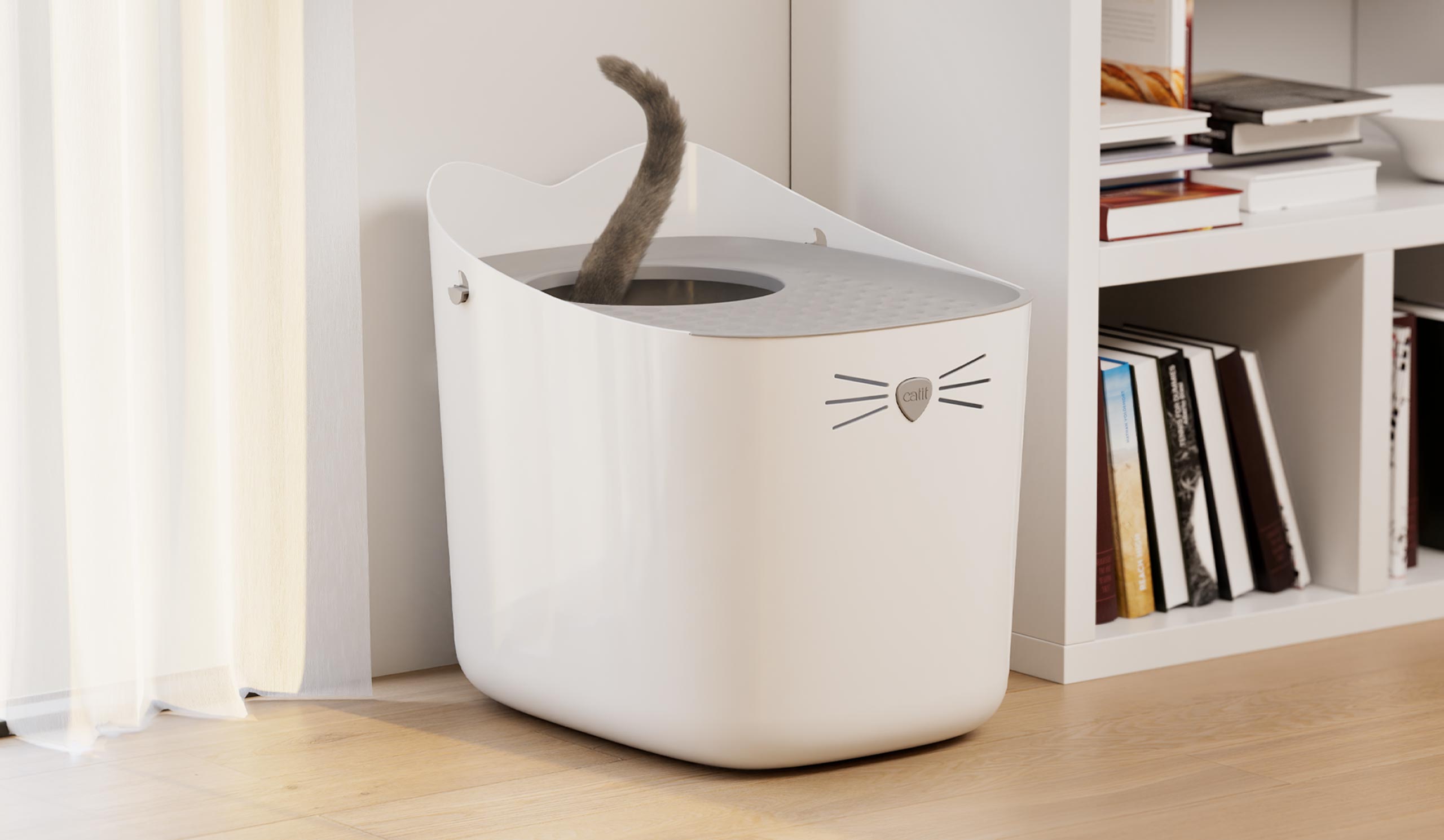 A white Catit PIXI litter box with a cat's tail visible, positioned on a wooden floor next to a bookshelf