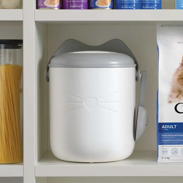 White cat-shaped food container with a grey lid and scoop, sitting on a shelf next to cat food packaging