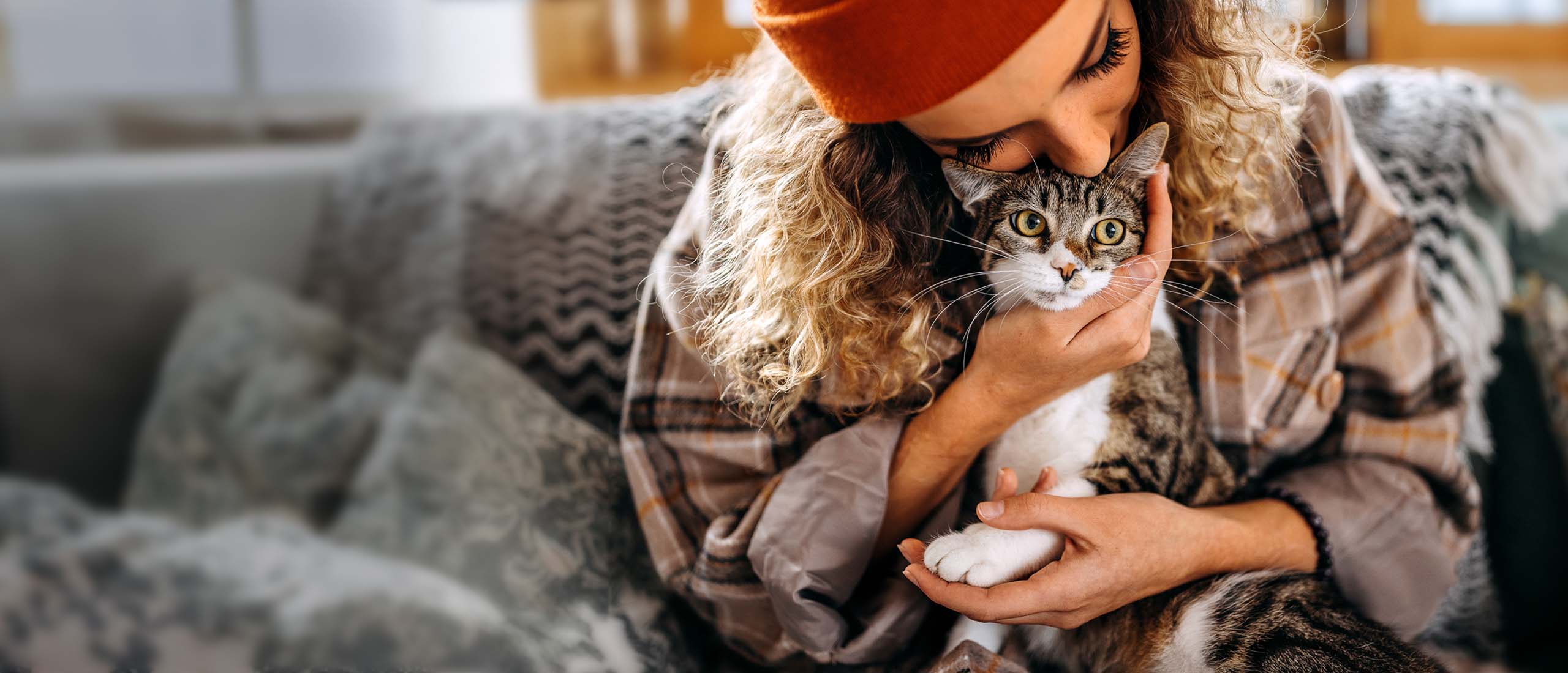A woman with curly hair wearing an orange beanie gently hugs and kisses a tabby cat on the head while sitting on a couch