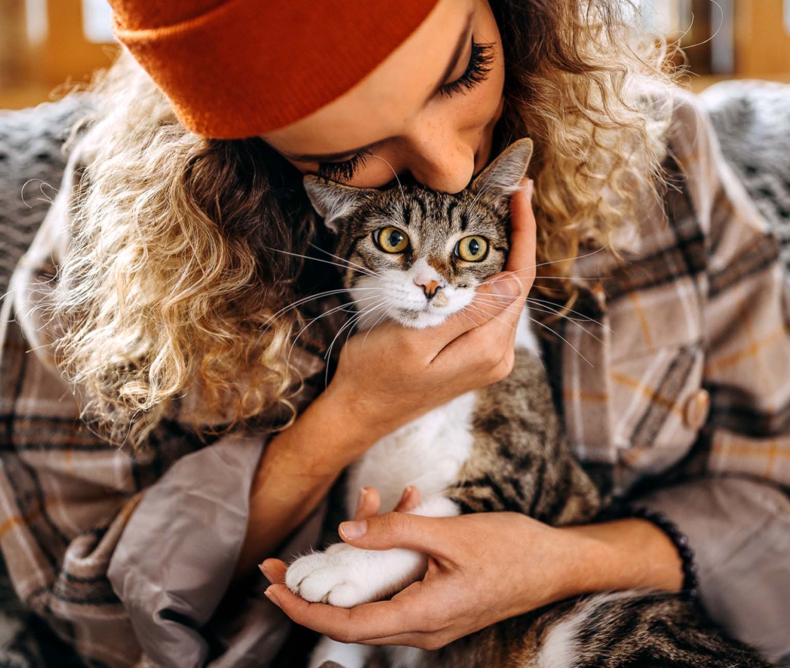 A woman wearing an orange beanie gently hugs and kisses a tabby cat while holding its paw
