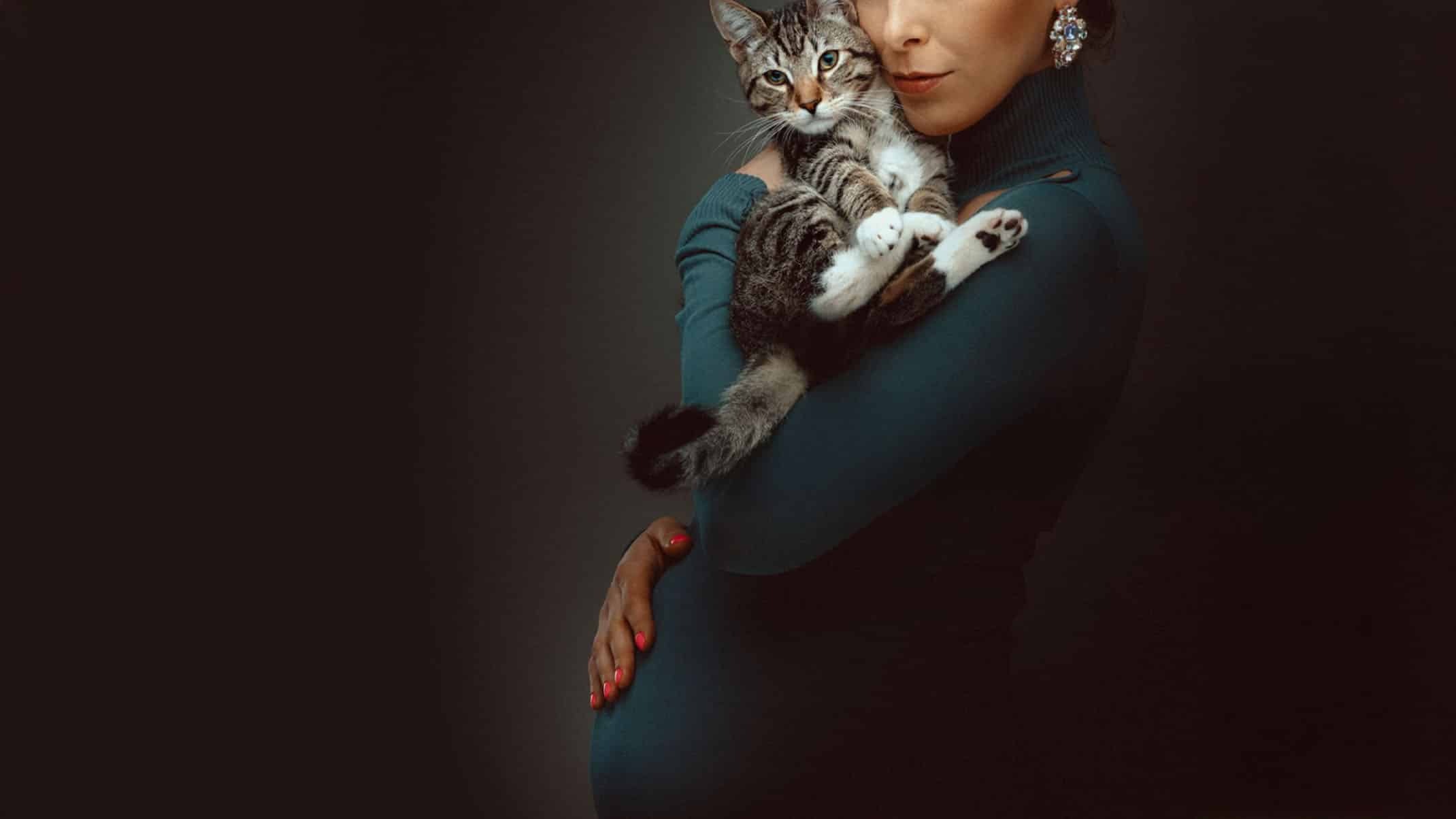 A pregnant woman in a teal turtleneck dress holding a tabby cat against a dark background