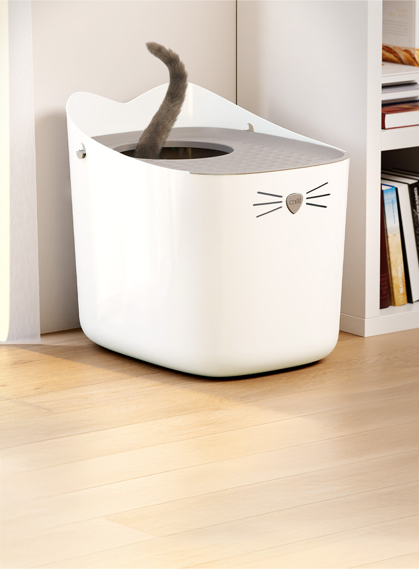 A white Catit PIXI litter box with a cat tail sticking out of the top opening, placed on a wooden floor next to a bookshelf
