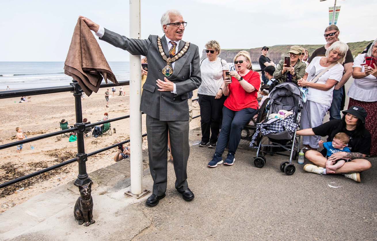 A man in a suit and ceremonial chain unveils a bronze cat statue on a seaside promenade while onlookers watch