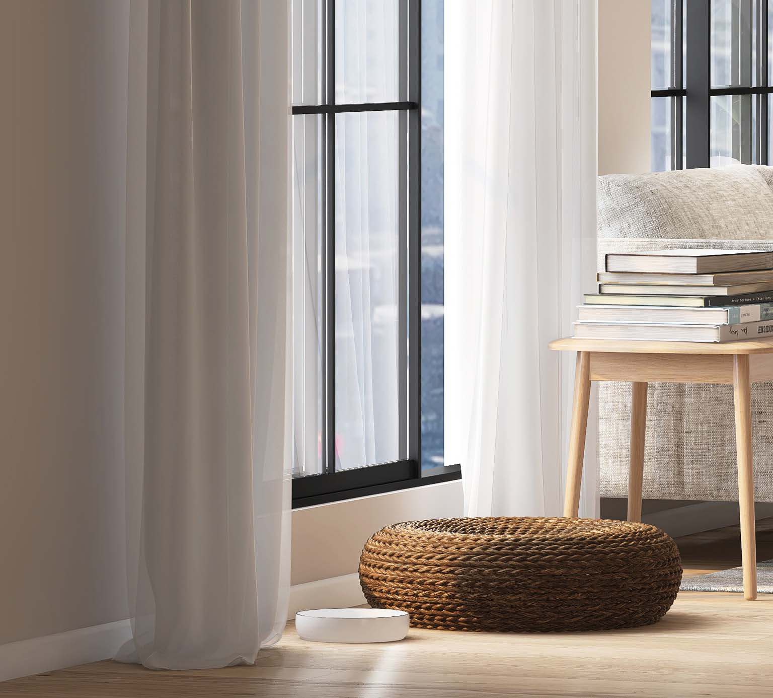 A white cat bowl sits on a wooden floor next to a woven pet bed near a window with sheer curtains