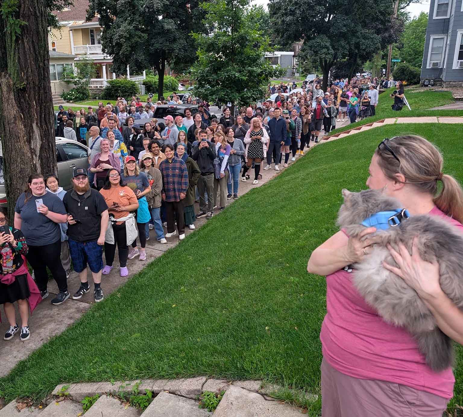 A woman holds a fluffy cat in a blue harness while a long line of people waits on a sidewalk in a residential neighborhood