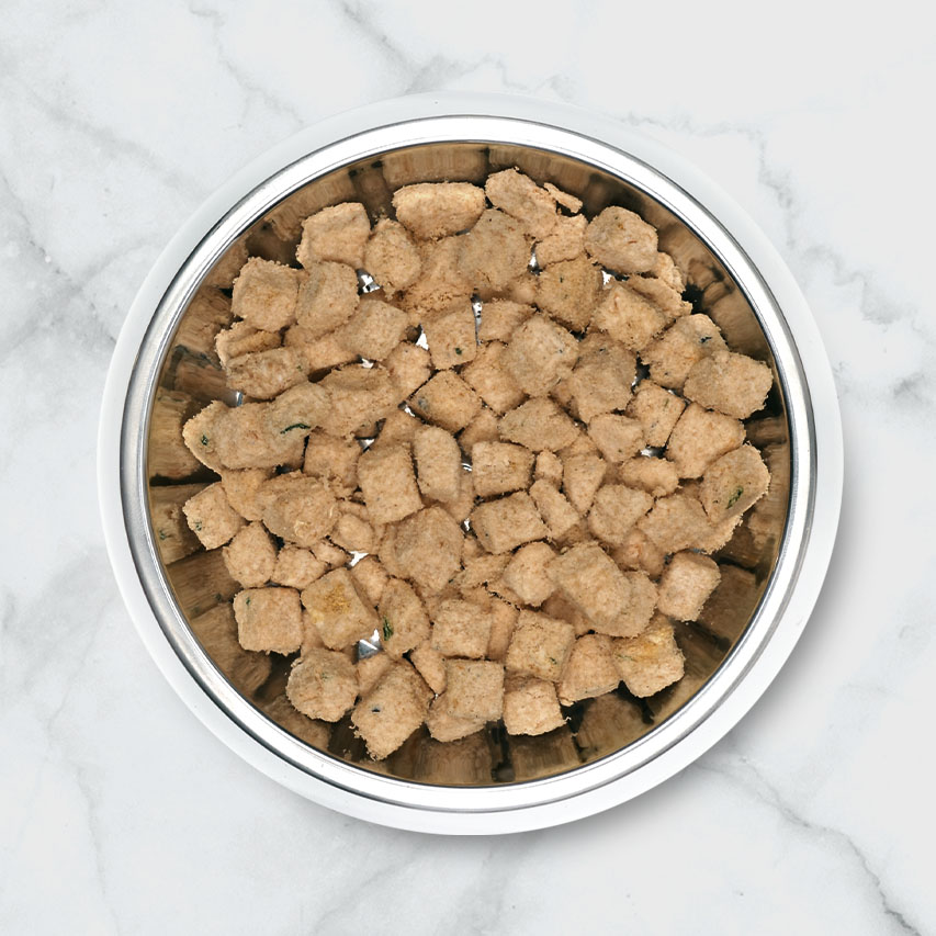 A metal bowl filled with small, square-shaped freeze-dried cat food pieces on a white marble surface