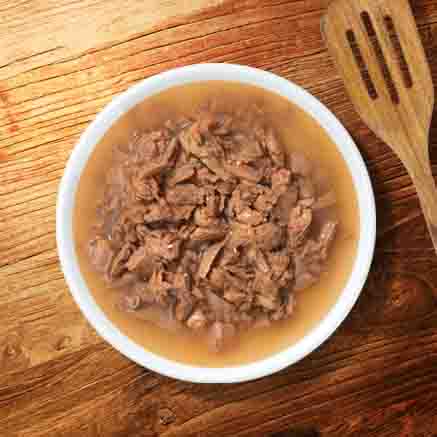 A white bowl filled with wet cat food in gravy, sitting on a wooden surface next to a wooden spatula