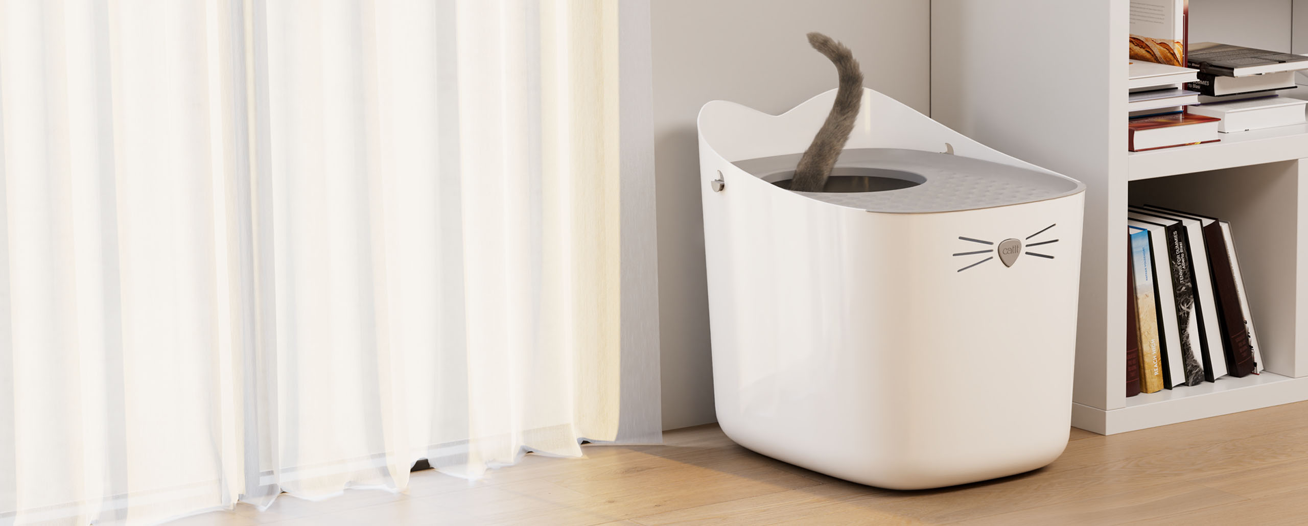 A white Catit PIXI litter box with a cat's tail visible inside, placed on a wooden floor next to a bookshelf