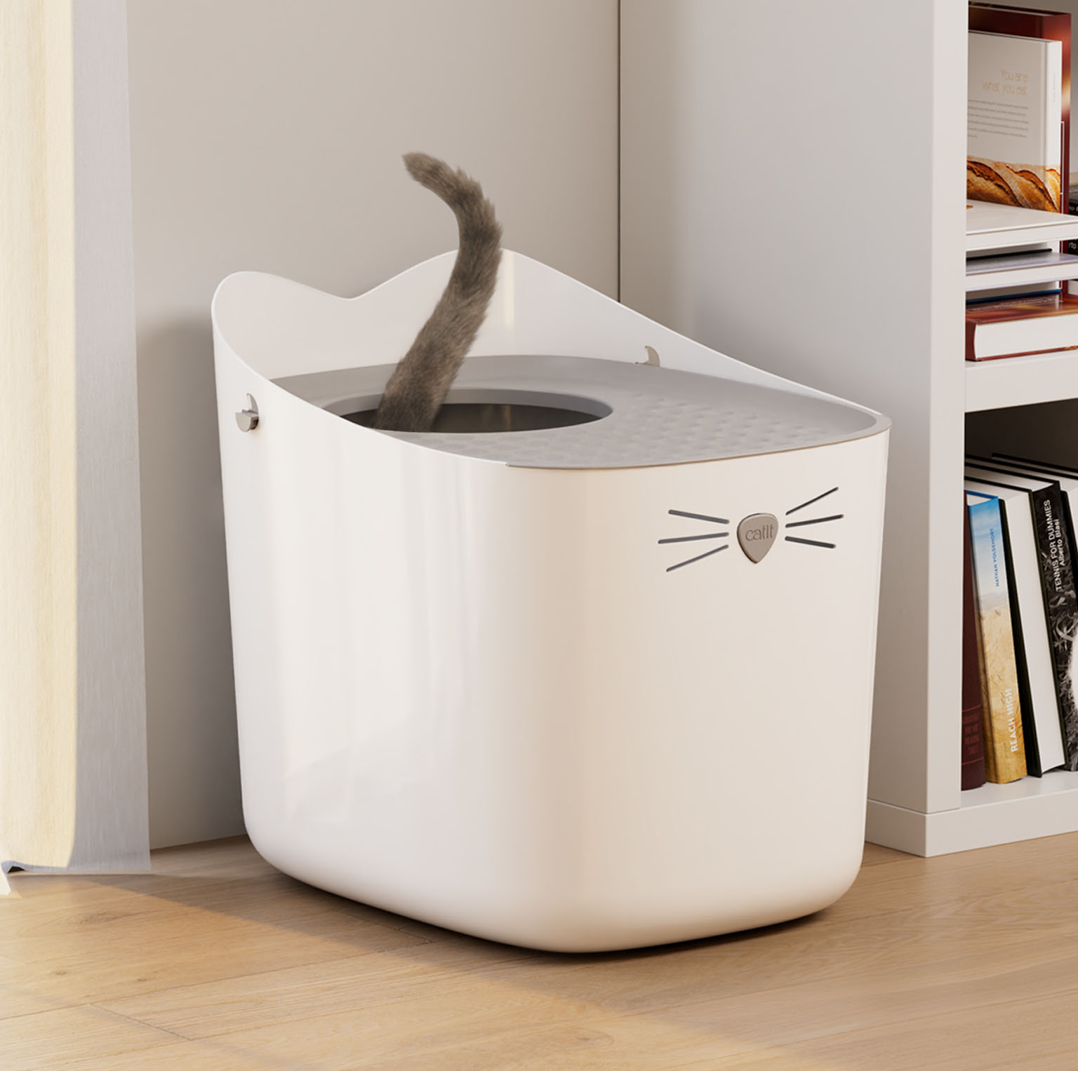 A white Catit PIXI litter box with a cat tail visible inside, positioned next to a bookshelf