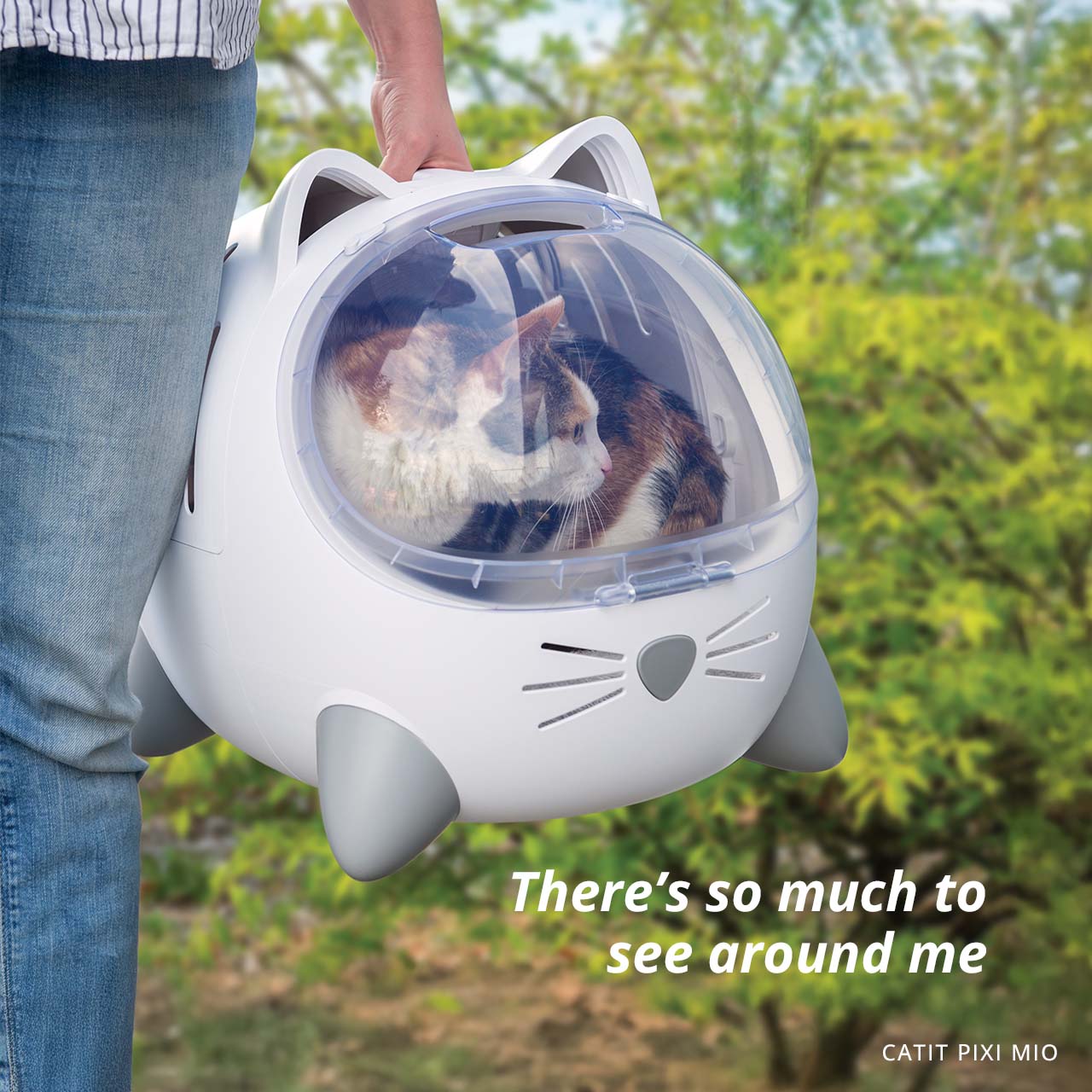A person carries a white, cat-shaped PIXI MIO pet carrier with a calico cat inside, outdoors