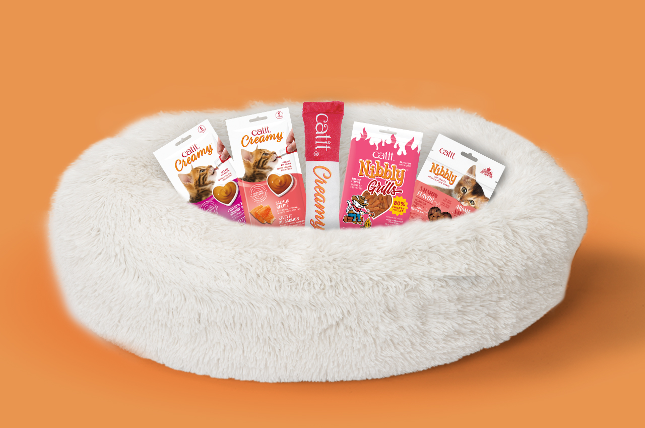 Catit Creamy and Nibbly cat treats arranged in a plush white cat bed against an orange background