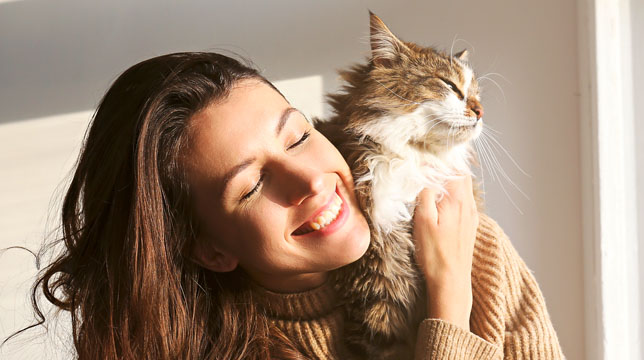 A woman smiling while holding and cuddling a fluffy tabby cat in the sunlight