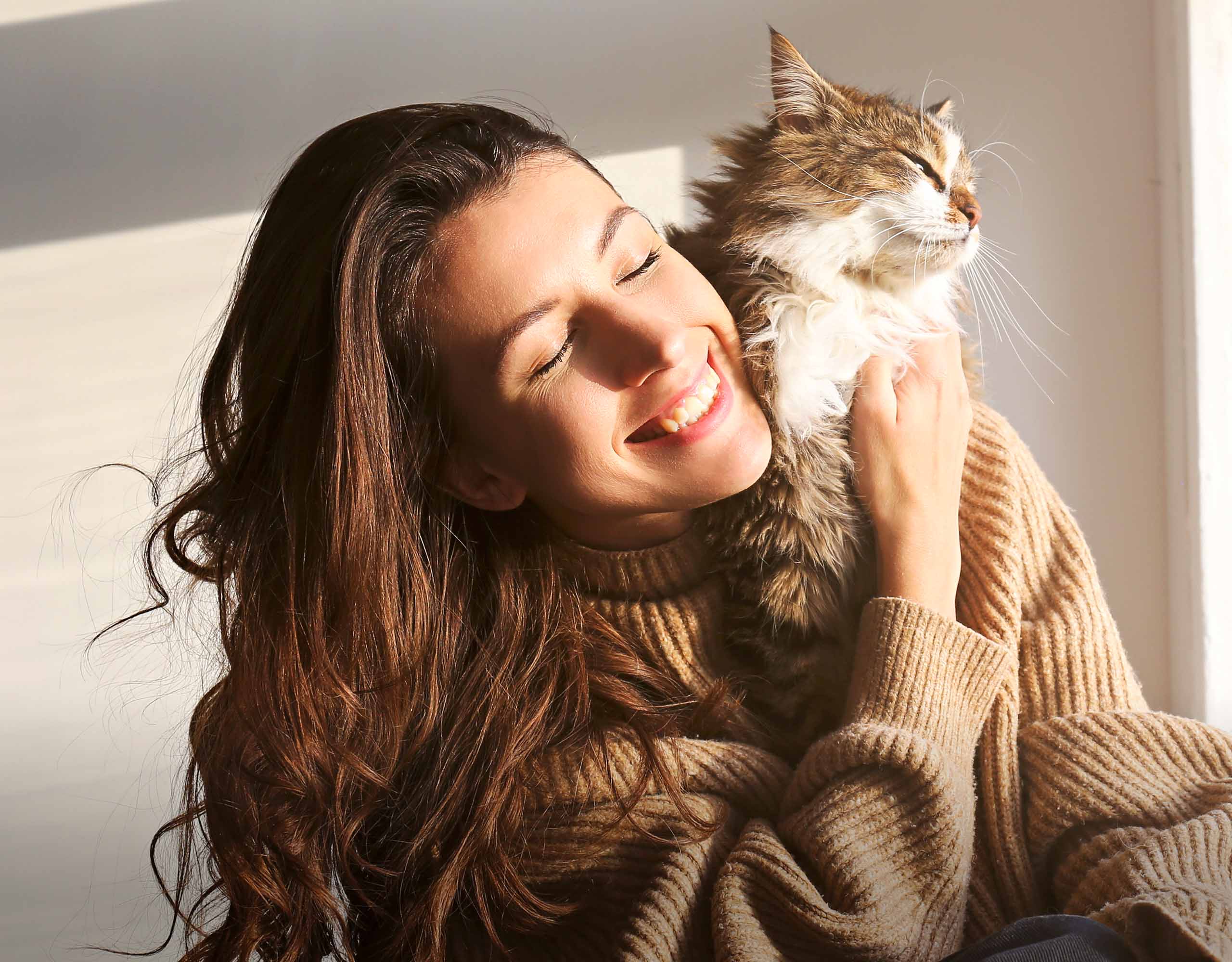 A woman smiling while hugging a fluffy brown tabby cat in a sunlit room