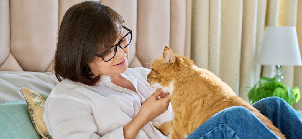 A woman wearing glasses and a white shirt sits on a bed, gently petting an orange cat resting on her lap