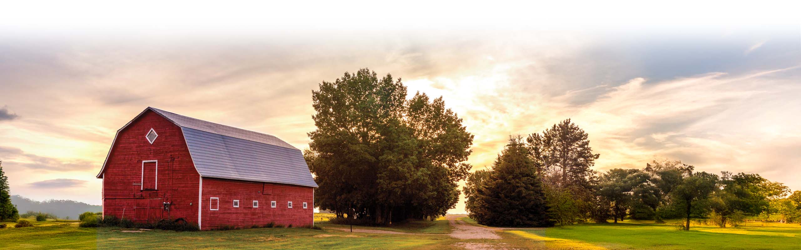 A red barn sits in a grassy field under a sunset sky