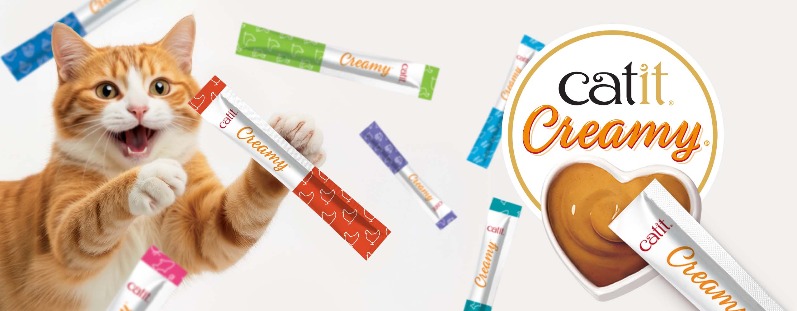An excited ginger cat holding a Catit Creamy treat stick, surrounded by various flavors of the treat and a heart-shaped bowl