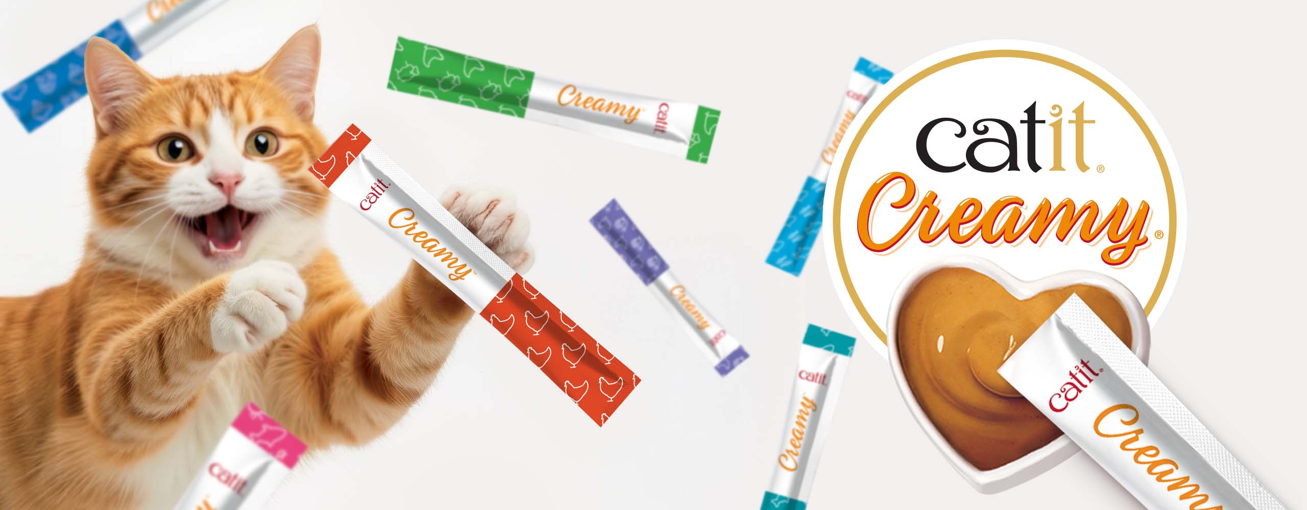 An orange tabby cat excitedly reaching for a Catit Creamy treat tube, surrounded by various flavors of the treat packaging