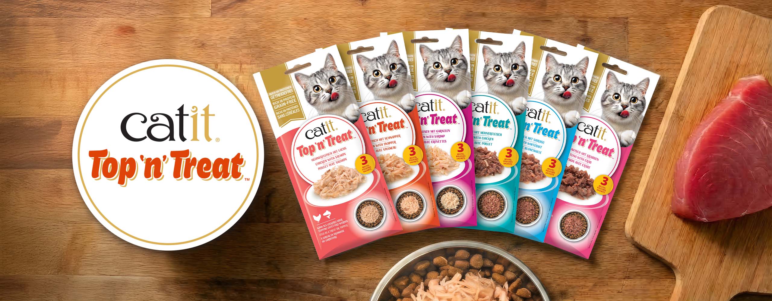 Catit Top 'n' Treat cat food pouches in six different flavors arranged on a wooden surface with a piece of raw fish