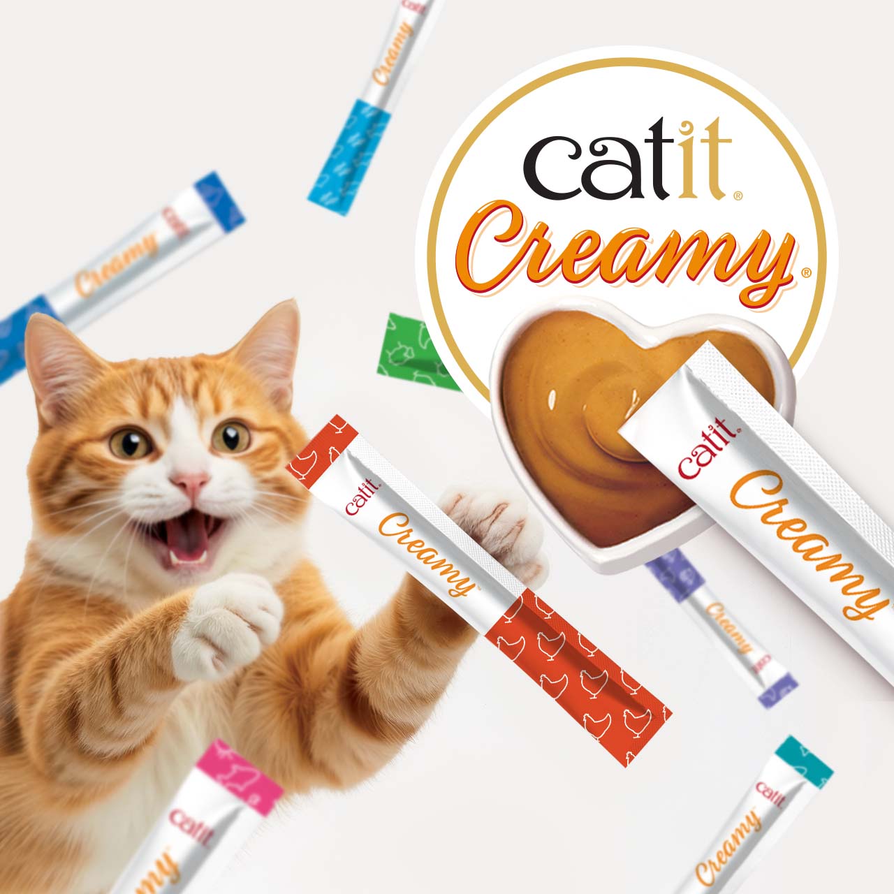 An orange tabby cat reaching for Catit Creamy cat treat sticks surrounding a heart-shaped bowl of creamy treat