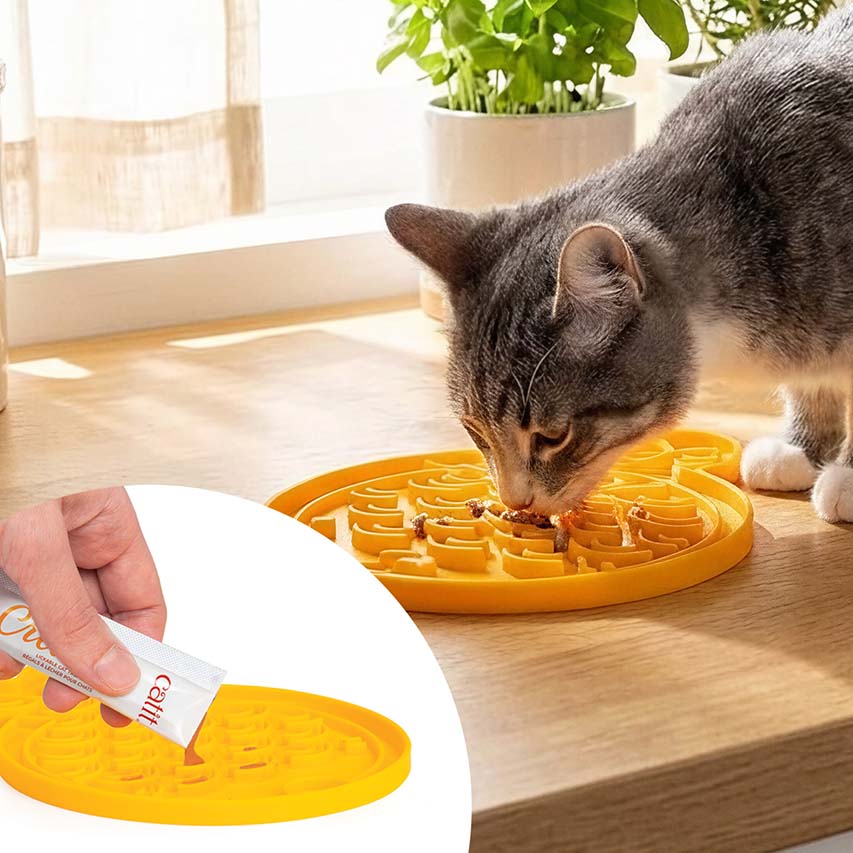 A cat eating from a yellow Catit lickable treat mat, with an inset showing a hand squeezing a Catit treat tube onto the mat