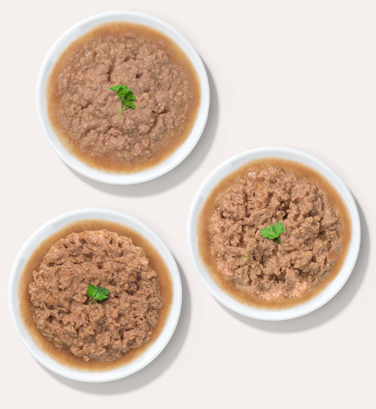 Three white bowls filled with wet cat food, each garnished with a small sprig of parsley