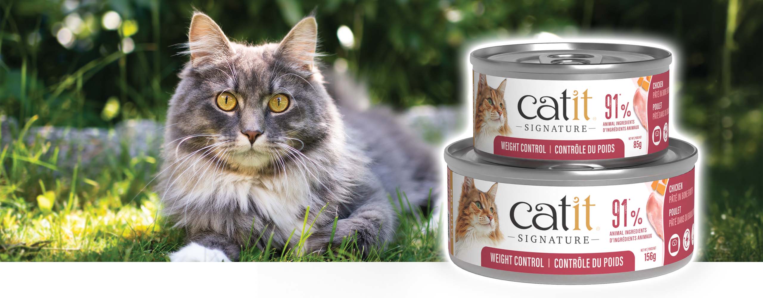 A fluffy grey cat lying in the grass next to two cans of Catit Signature Weight Control cat food