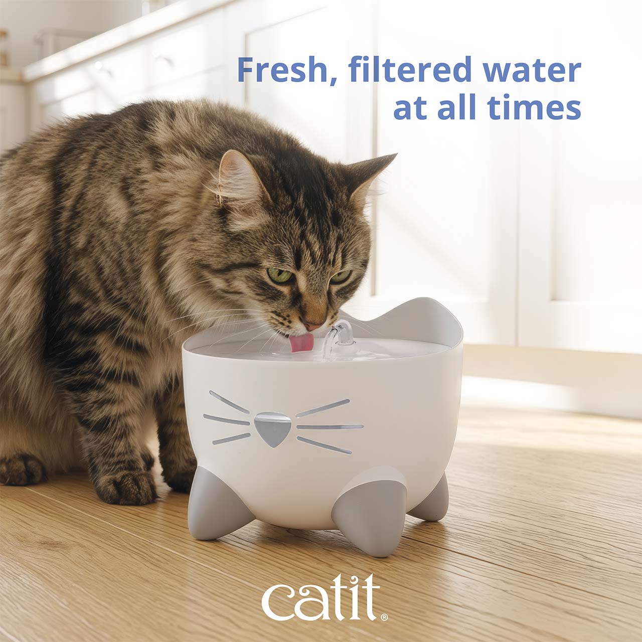 A fluffy tabby cat drinks from a white Catit PIXI cat water fountain shaped like a cat face