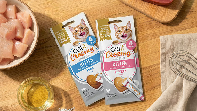 Two packages of Catit Creamy Booster kitten treats in tuna and chicken flavors, displayed on a wooden surface with ingredients