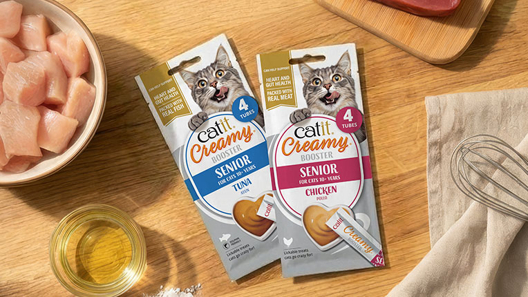 Two packages of Catit Creamy Booster treats for senior cats, one tuna and one chicken flavor, on a wooden surface with ingredients