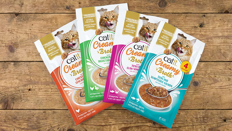 Four packages of Catit Creamy Broth cat treats in various flavors arranged on a wooden surface