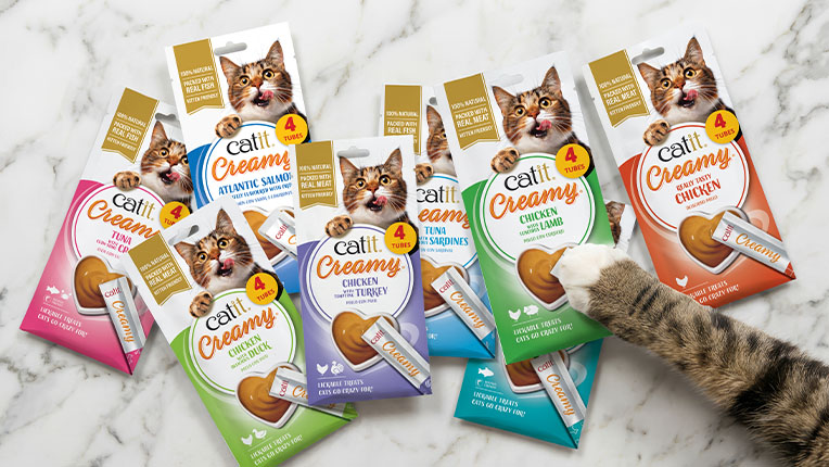 A cat's paw reaches toward several packages of Catit Creamy lickable cat treats in various flavors