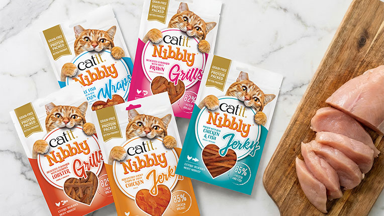 Five packages of Catit Nibbly cat treats arranged around raw chicken breast slices on a wooden cutting board