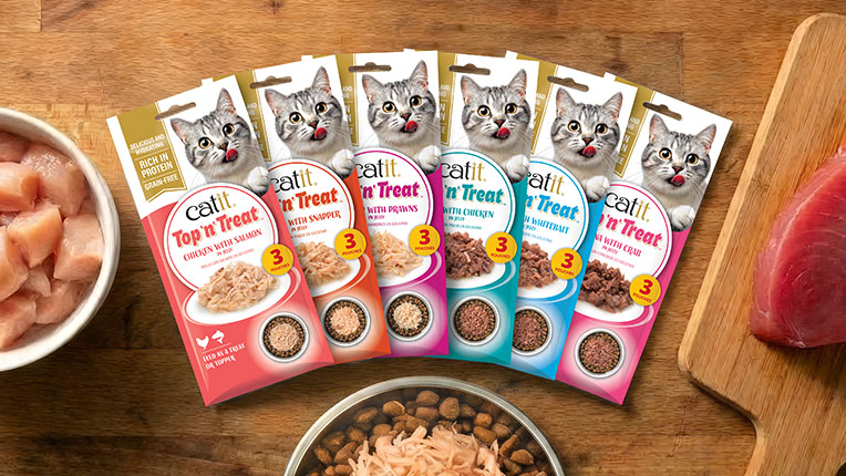 Six varieties of Catit Top 'n' Treat cat food pouches arranged in a fan shape on a wooden surface with raw chicken and tuna nearby