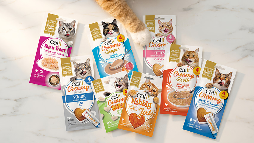 A variety of Catit cat treat packaging arranged on a marble surface with a cat's paw reaching into the frame